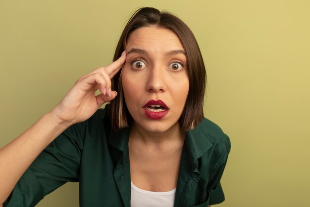 Mujer bonita ansiosa pone los dedos en la sien y mira al frente aislado en la pared verde oliva