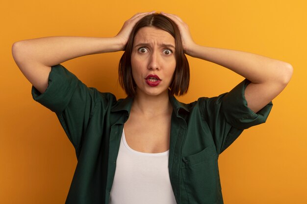 Mujer bonita ansiosa pone las manos en la cabeza aislada en la pared naranja