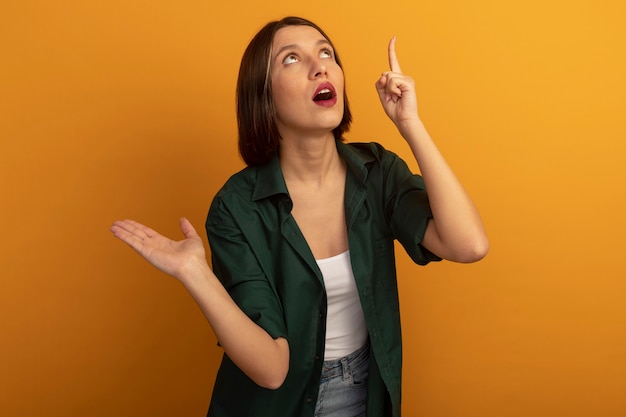 Mujer bonita ansiosa sostiene la mano abierta mirando y apuntando hacia arriba aislado en la pared naranja