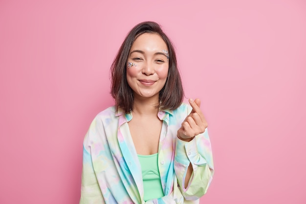 Mujer bonita asiática hace coreano como signo mini escuchar gesto chasquear los dedos tiene cabello oscuro natural vestido con camisa colorida aislada sobre pared rosa expresa amor. Concepto de lenguaje corporal.