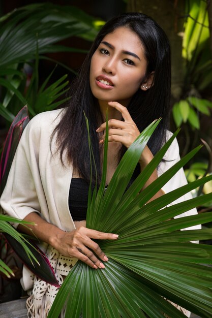 Mujer bonita asiática posando en el jardín tropical, sosteniendo una gran hoja de palma.