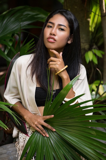 Mujer bonita asiática posando en el jardín tropical, sosteniendo una gran hoja de palma.