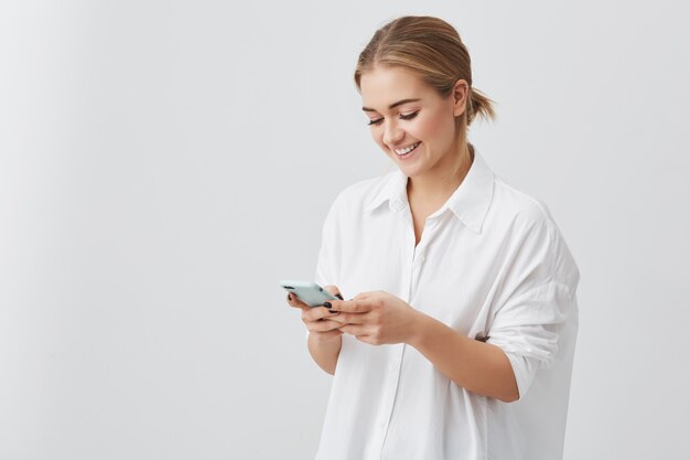 Mujer bonita atractiva con el pelo rubio en camisa blanca sonriendo mientras usa el teléfono celular chateando con su novio posando. Concepto de belleza y juventud