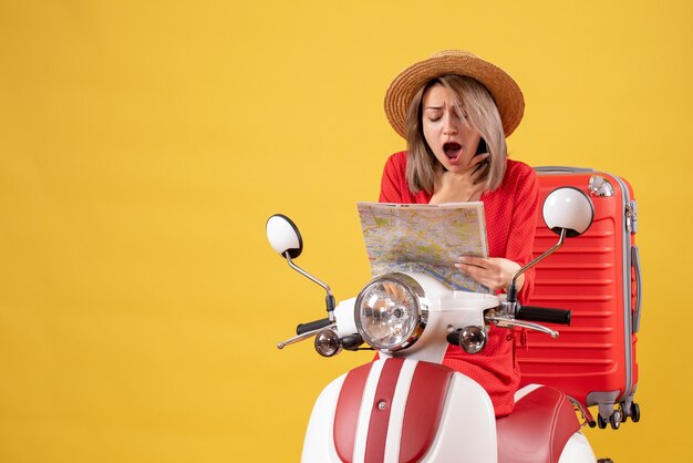 Mujer bonita aturdida en ciclomotor con maleta roja sosteniendo el mapa