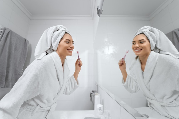 Mujer bonita en bata blanca con toalla en la cabeza, cepillarse los dientes en el baño.