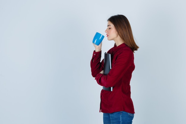 Mujer bonita bebiendo té, sosteniendo la carpeta en blusa burdeos y buscando sed. .