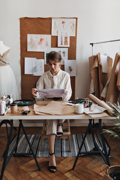 Mujer bonita en blusa blanca mira la mesa de encaje Dama morena en camisa beige y pantalones posando en la oficina del diseñador