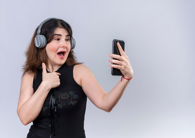 Mujer bonita en blusa negra escuchando música con auriculares haciendo slefie con teléfono