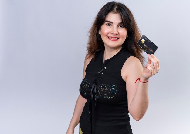 Mujer bonita en blusa negra mostrando tarjeta de crédito mira felizmente a cámara
