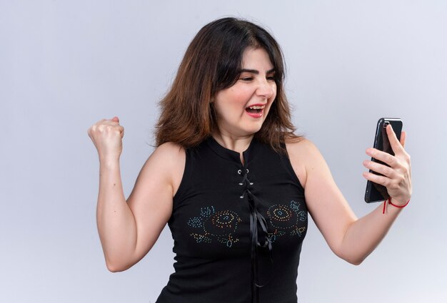Mujer bonita en blusa negra sosteniendo el teléfono y mirando feliz por ganar