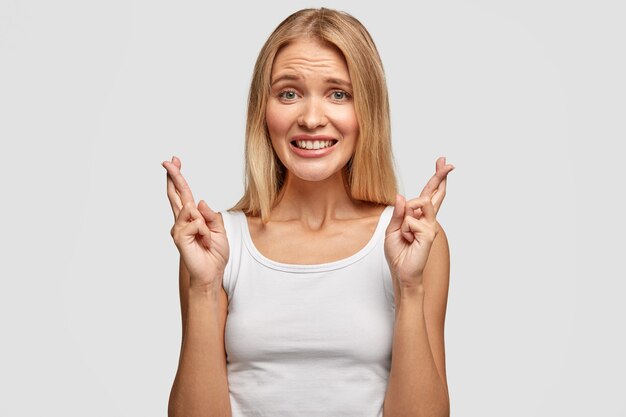 Mujer bonita con cabello claro, cruza los dedos, cree en la buena suerte, tiene una sonrisa con dientes, vestida con ropa casual