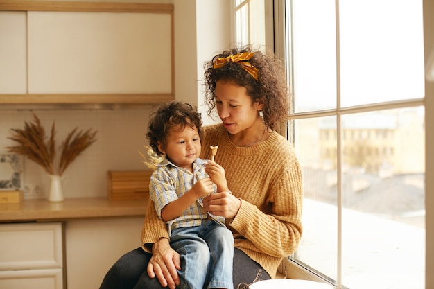 Mujer bonita con cabello rizado sentada en el alféizar de la ventana con un bebé adorable en su regazo, dándole juguetes o dulces, niño mirando con interés y curiosidad. Maternidad, puericultura y convivencia