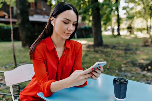 Mujer bonita con camisa roja sentada en la mesa con teléfono inteligente y escuchando música