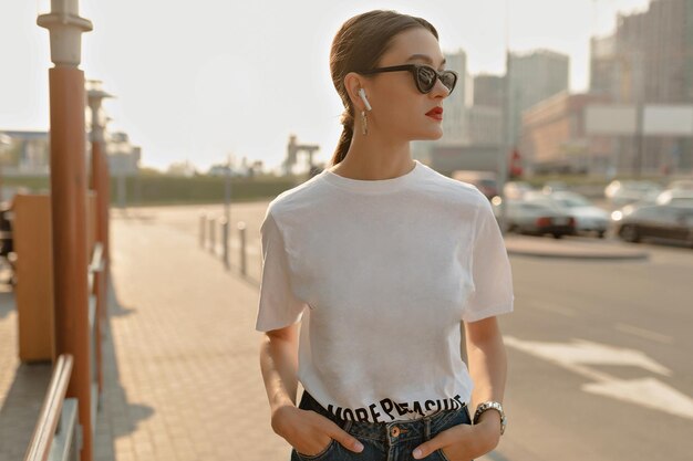 Mujer bonita en camiseta y jeans y gafas de sol posando en el fondo de la ciudad Bella dama en gafas oscuras con cabello oscuro y labios rojos camina afuera