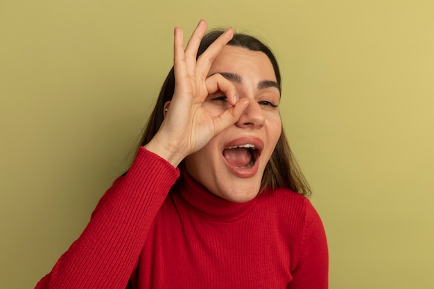 Foto gratuita mujer bonita caucásica alegre gestos ok signo de la mano y mira a la cámara a través de los dedos en verde oliva