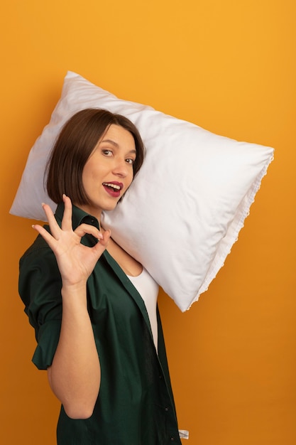 Foto gratuita mujer bonita caucásica complacida se para de lado sosteniendo la almohada en el hombro y gesticulando con la mano ok