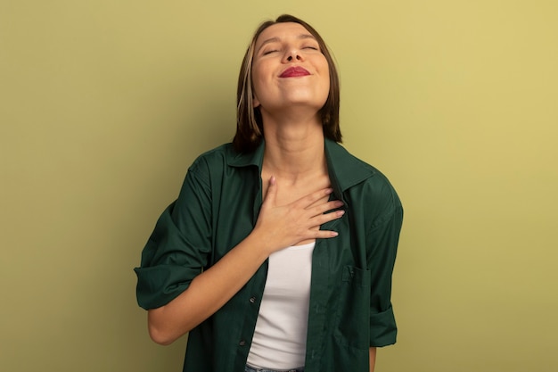 Mujer bonita caucásica complacida pone la mano en el pecho en verde oliva