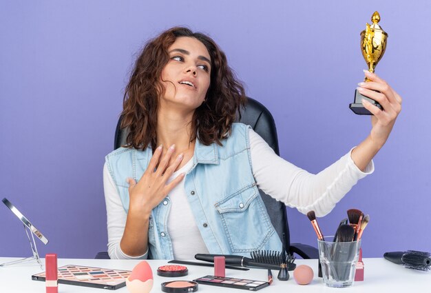 Mujer bonita caucásica complacida sentada en la mesa con herramientas de maquillaje sosteniendo y mirando la copa ganadora y poniendo la mano sobre su pecho