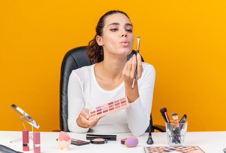 Foto gratuito mujer bonita caucásica complacida sentada en la mesa con herramientas de maquillaje sosteniendo la paleta de sombras de ojos y mirando pinceles de maquillaje aislados en la pared naranja con espacio de copia