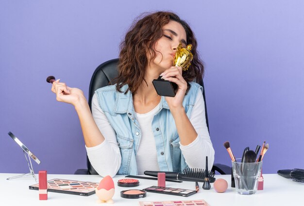 Mujer bonita caucásica complacida sentada a la mesa con herramientas de maquillaje sosteniendo un pincel de maquillaje y besando la copa ganadora aislada en la pared púrpura con espacio de copia