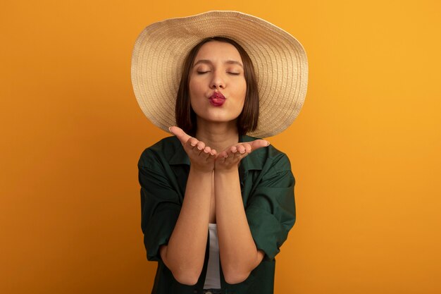 Mujer bonita caucásica complacida con sombrero de playa envía beso con dos manos en naranja