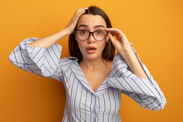 Mujer bonita caucásica sorprendida con la mano en la cabeza y mira a la cámara a través de gafas ópticas en naranja