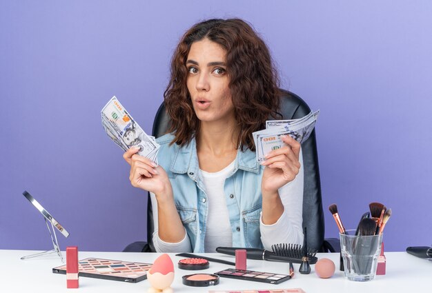 Mujer bonita caucásica sorprendida sentada en la mesa con herramientas de maquillaje con dinero