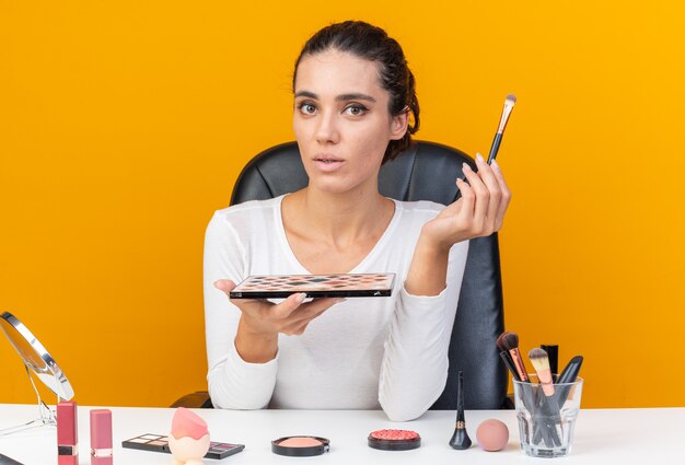 Mujer bonita caucásica sorprendida sentada en la mesa con herramientas de maquillaje con pincel de maquillaje y paleta de sombra de ojos