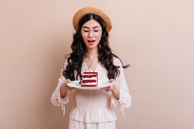 Mujer bonita China en vasos con plato con postre. Atractiva mujer asiática rizada mirando pastel.