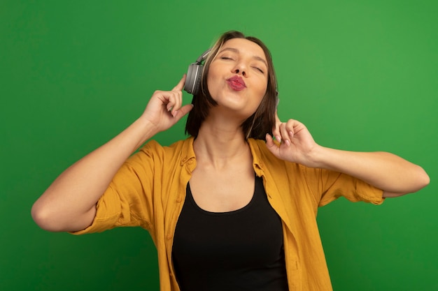 Mujer bonita complacida en auriculares finge besar aislado en la pared verde