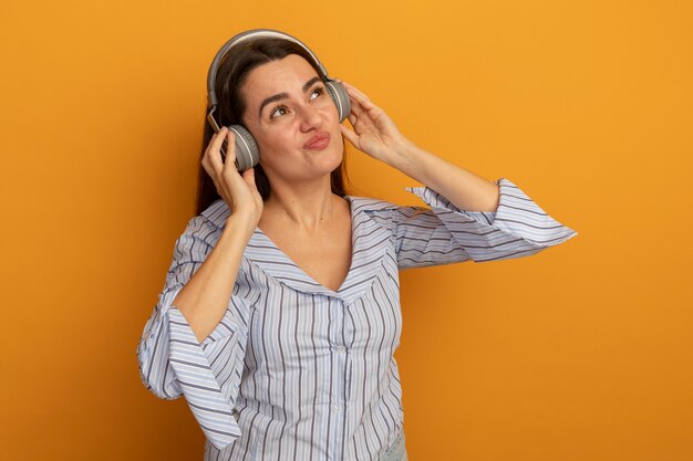 Mujer bonita complacida en auriculares mira al lado aislado en la pared naranja