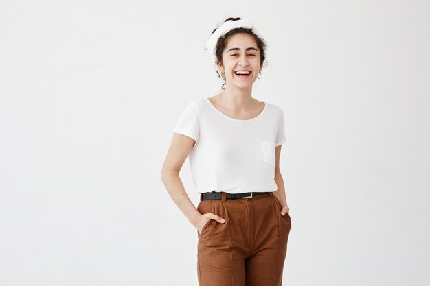 Mujer bonita complacida con cabello oscuro y ondulado en moño, ojos oscuros y piel sana vestida con una camiseta blanca y pantalones marrones tomados de las manos en los bolsillos, posando contra la pared blanca.