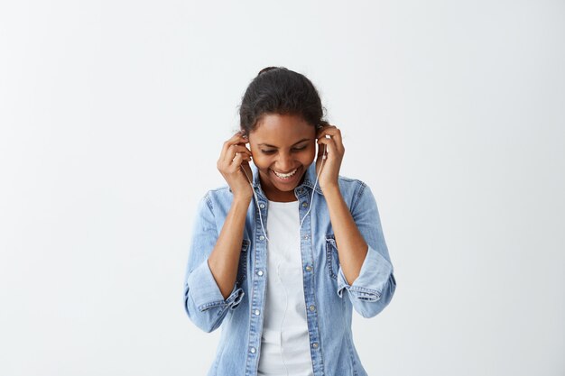 Mujer bonita complacida con moño, ojos oscuros y piel oscura sana vestida con una camiseta blanca, chaqueta azul, con los auriculares puestos, sonriendo mientras posa contra el muro de hormigón blanco. Personas y estilo de vida.