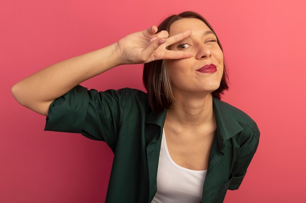 Mujer bonita complacida parpadea y gesticula el signo de la mano de la victoria mirando al frente a través de los dedos aislados en la pared rosa