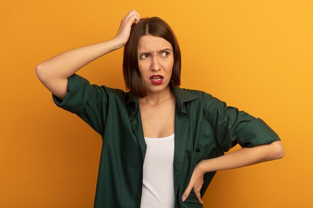 Mujer bonita despistada pone la mano en la cabeza y mira el lado aislado en la pared naranja