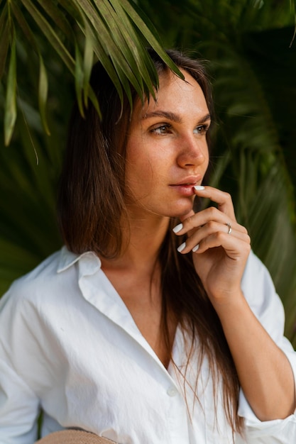 Mujer bonita disfrutando de unas vacaciones tropicales en Tailandia, vistiendo una blusa blanca y posando sobre palmeras.