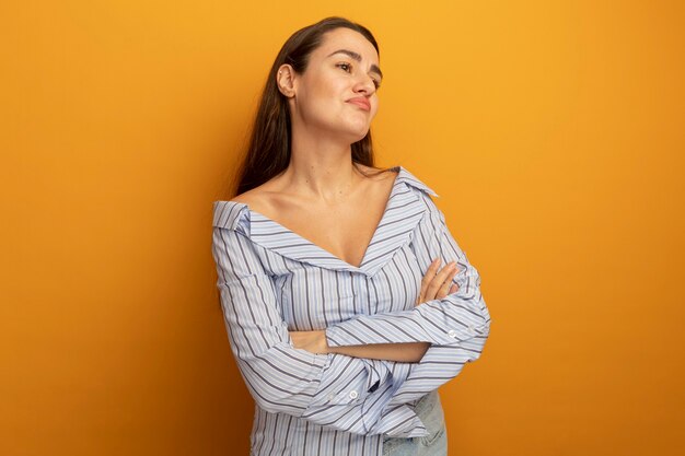 Mujer bonita disgustada se encuentra con los brazos cruzados mirando al lado aislado en la pared naranja