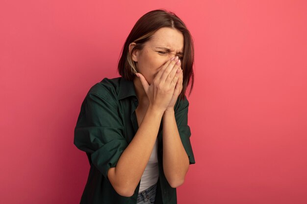 Mujer bonita disgustada pone las manos en la nariz aislada en la pared rosa