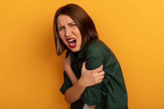 Mujer bonita dolorida sostiene los brazos y grita aislado en la pared naranja