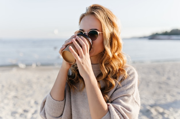 Mujer bonita en elegantes gafas de sol descansando en la costa del mar. Retrato al aire libre de modelo femenino de ensueño escalofriante en día de otoño.