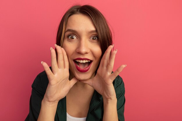 Mujer bonita emocionada tiene las manos cerca de la boca mirando al frente aislado en la pared rosa