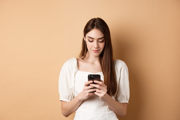 Mujer bonita enviando mensajes de texto en la pantalla de lectura del teléfono celular del teléfono inteligente y sonriendo de pie sobre fondo beige