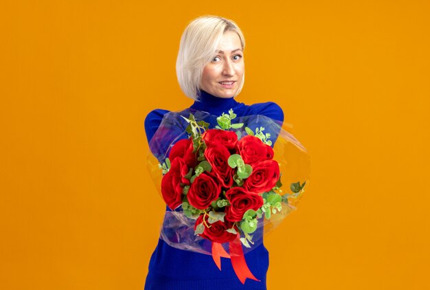 Mujer bonita eslava sonriente sosteniendo un ramo de flores en el día de San Valentín aislado en la pared naranja con espacio de copia