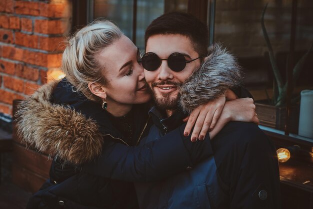 La mujer bonita está besando a su hombre sonriente con gafas de sol cerca del café en el frío día de otoño.
