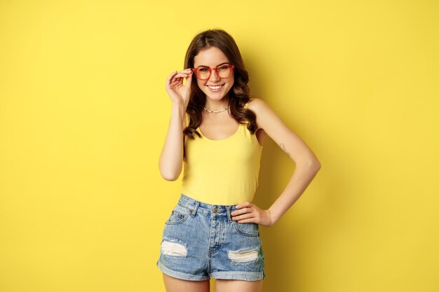 Mujer bonita con estilo posando con gafas de sol y ropa de verano, luciendo feliz y despreocupada, concepto de vacaciones y días soleados, de pie sobre fondo amarillo