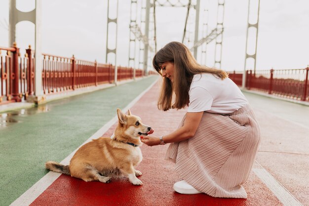 Mujer bonita con estilo vistiendo falda y camiseta blanca jugando con perro corgi en el puente soleado