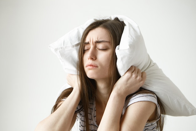 Mujer bonita estudiante en camiseta a rayas tratando de dormir un poco, sentada en el dormitorio con la almohada en la cabeza, cerrando los ojos