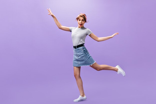 Mujer bonita en falda y camisa salta sobre fondo morado. Chica joven divertida con peinado elegante en zapatillas blancas posando.