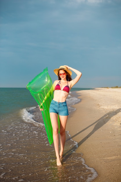 Foto gratuita mujer bonita feliz divirtiéndose en la playa, vistiendo ropa de playa elegante, sombrero de bikini y pantalones cortos de mezclilla, piernas largas, cuerpo delgado, sosteniendo colchón de aire y caminando cerca del océano.