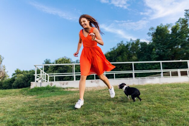 Mujer bonita feliz en el parque corriendo con el perro boston terrier, sonriente estado de ánimo positivo, estilo de moda de verano, vestido naranja, jugando con la mascota, divirtiéndose, coloridas y activas vacaciones de fin de semana, zapatillas de deporte